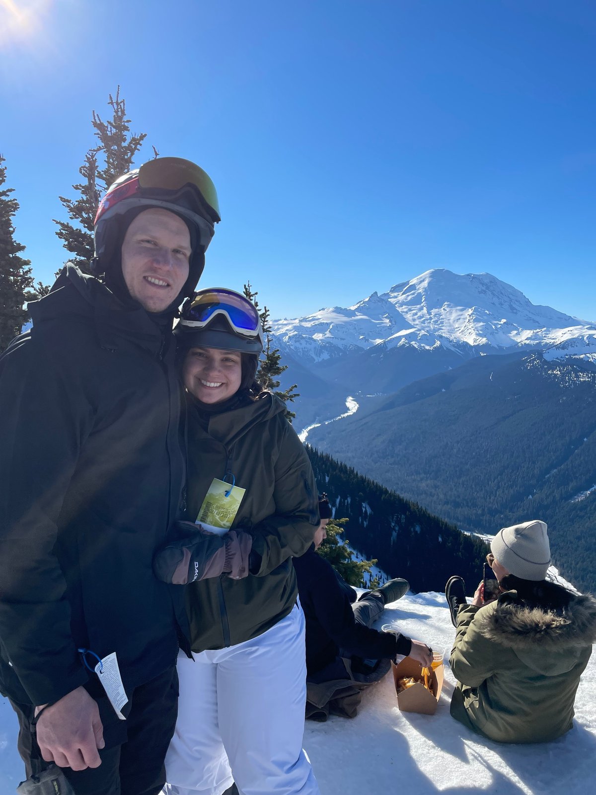 Two skiers smile at the camera on a snowy mountain with a snow-capped peak visible in the background