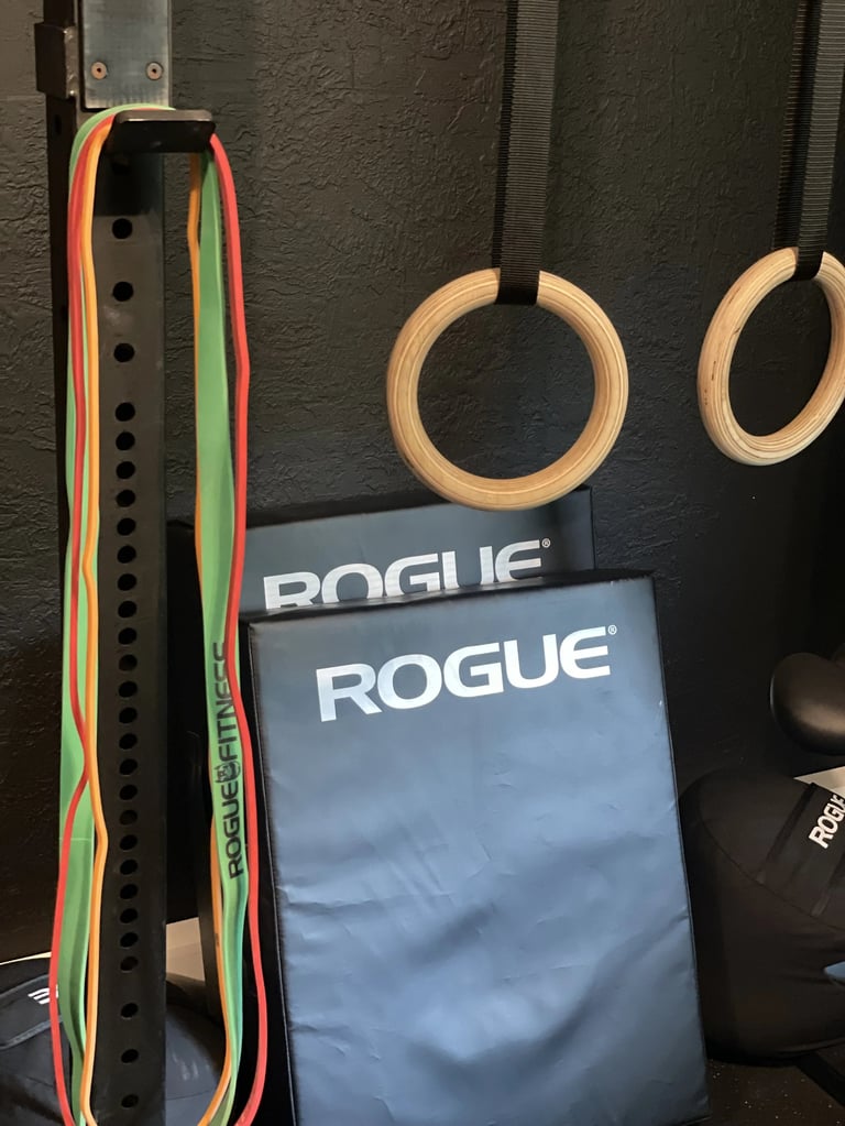 Gym equipment mounted on dark wall including wooden gymnastic rings, resistance bands, and Rogue branded items