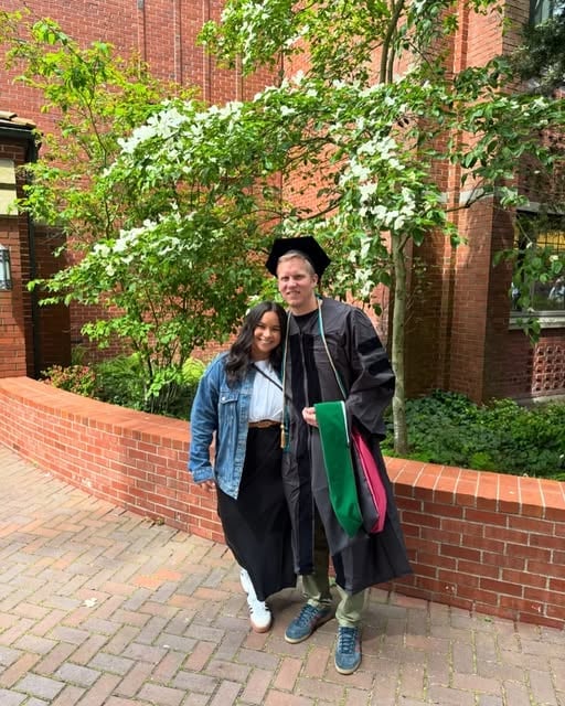Two people posing together in front of a brick building with ivy-covered archway, wearing casual clothing